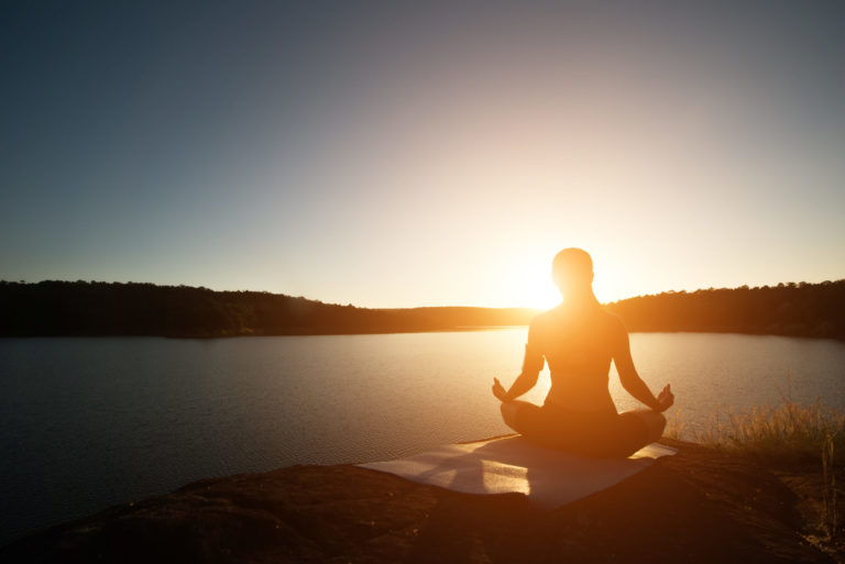 pessoa numa montanha meditando sentada, ao fundo o pôr do sol