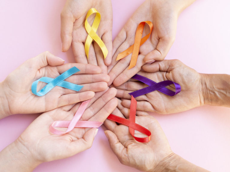 várias mãos lado a lado, cada mão possui um laço de fita com cor diferente, representando os meses de combate a cada tipo de câncer
